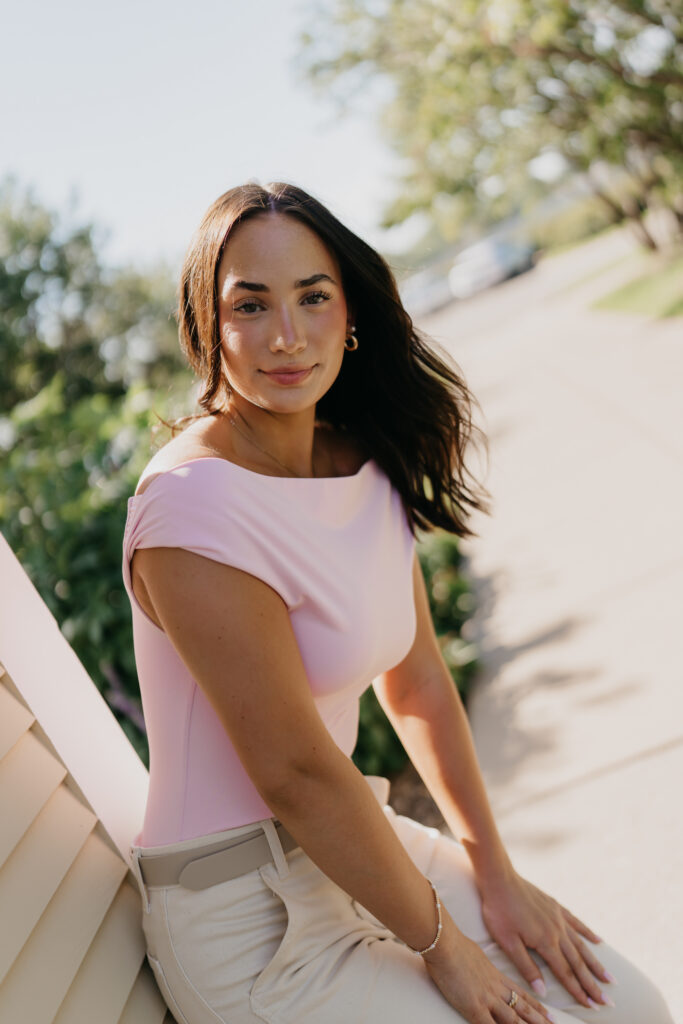 Senior girl in pink top and cream pants sitting on a stone patio ledge