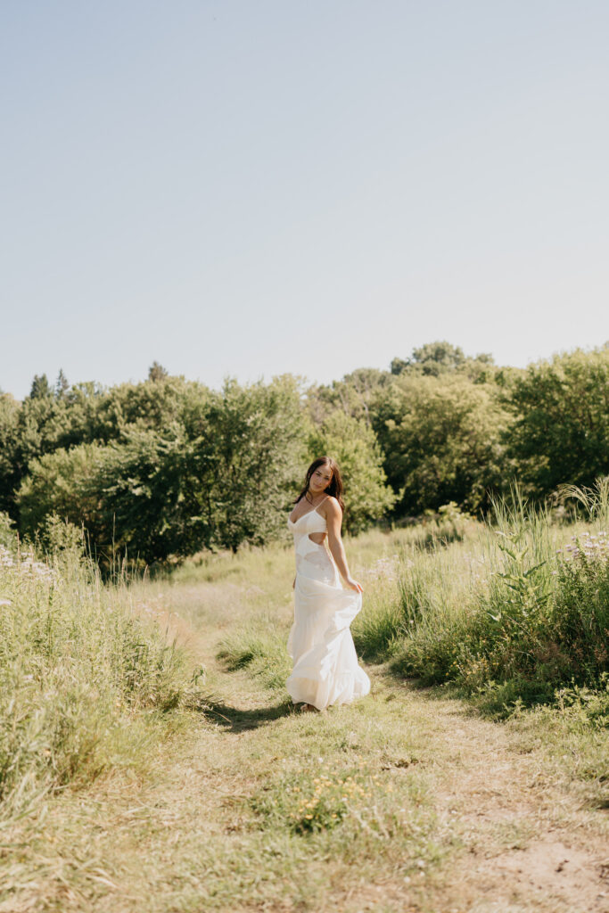 Senior girl in cream cut-out dress standing in a sunlit wildflower field