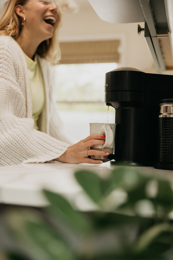Woman in a white knit sweater dress laughing while making coffee with an espresso machine