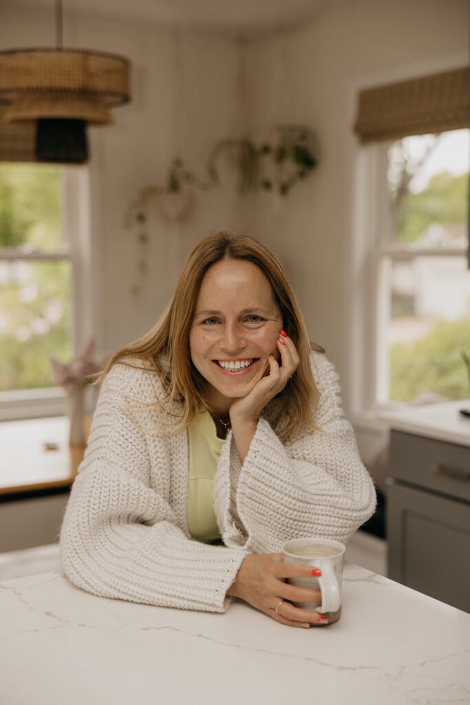 Woman in a white knit sweater dress sitting at a kitchen table smiling, resting her chin on her hand