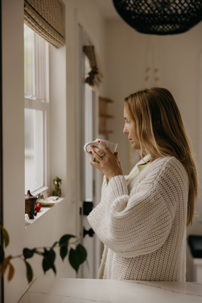 Woman in a white knit sweater dress drinking coffee by a sunlit kitchen window