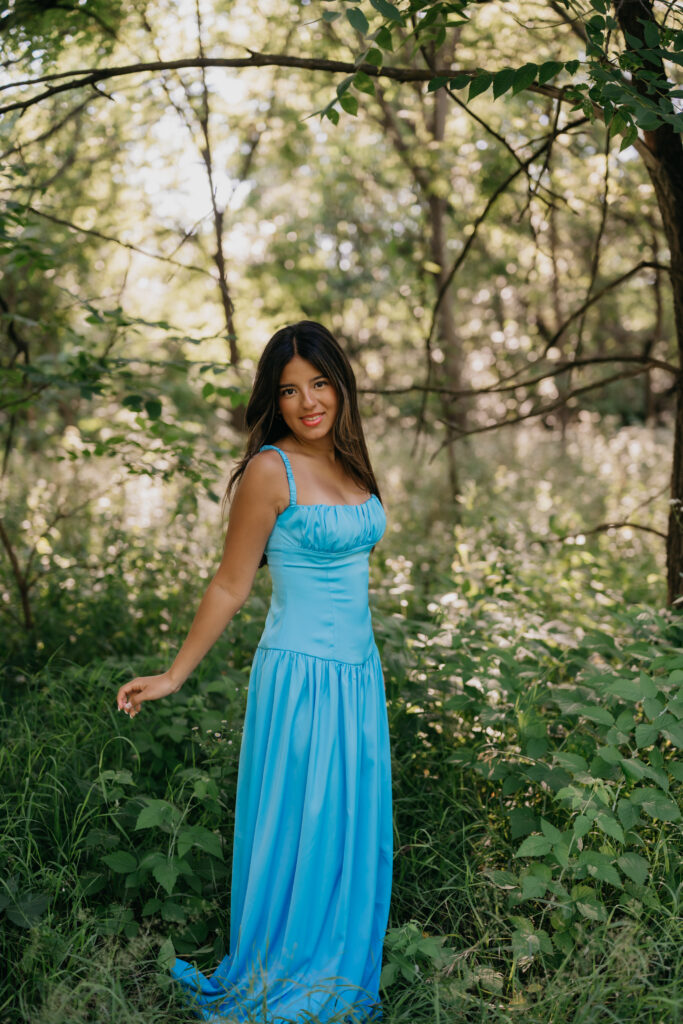 Natalia standing in a blue satin dress surrounded by greenery in the woods