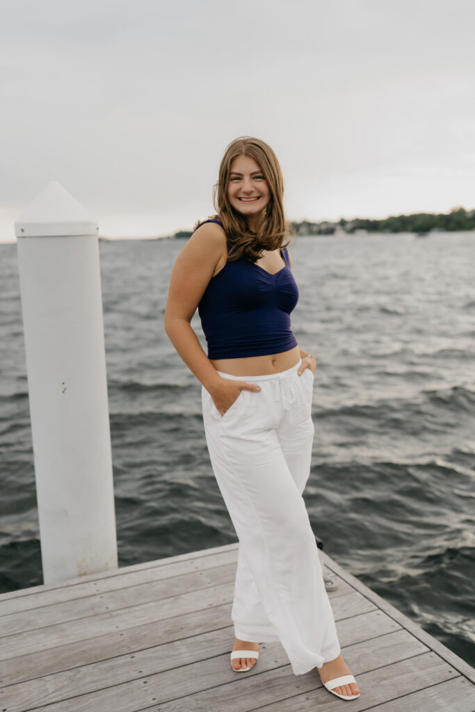 Senior Rachel standing on Wayzata Docks with her hand in her pocket, in a navy top and white pants
