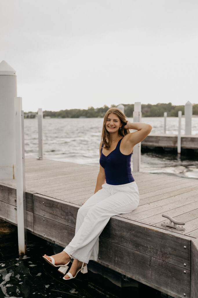 Rachel sitting on the edge of the dock in a navy top and white pants, legs crossed