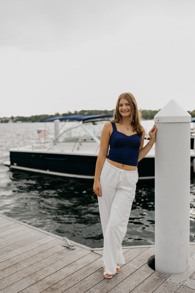 Senior Rachel standing on Wayzata Docks beside a piling, in a navy top and white pants