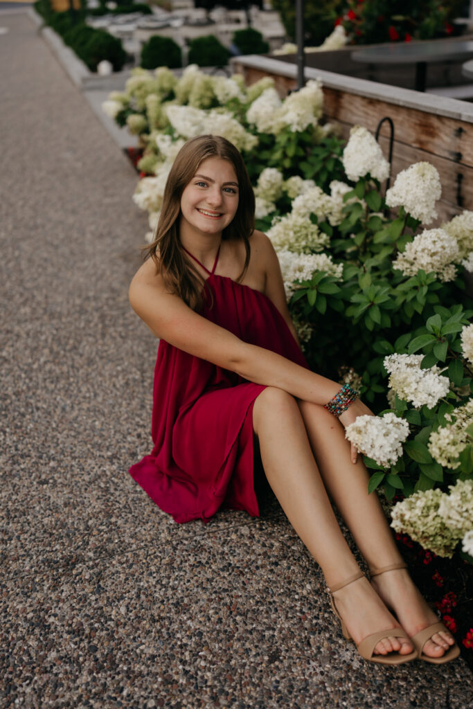 Rachel sitting beside white hydrangeas on a gravel path, in a red dress