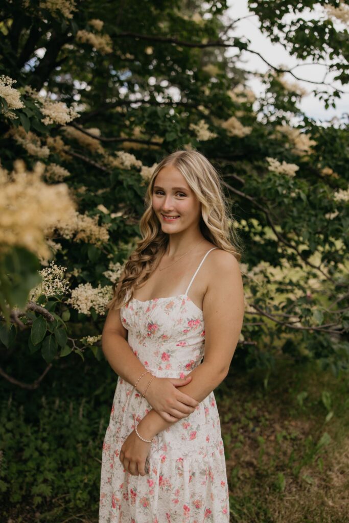 Senior girl in a floral maxi dress standing beneath flowering greenery smiling softly