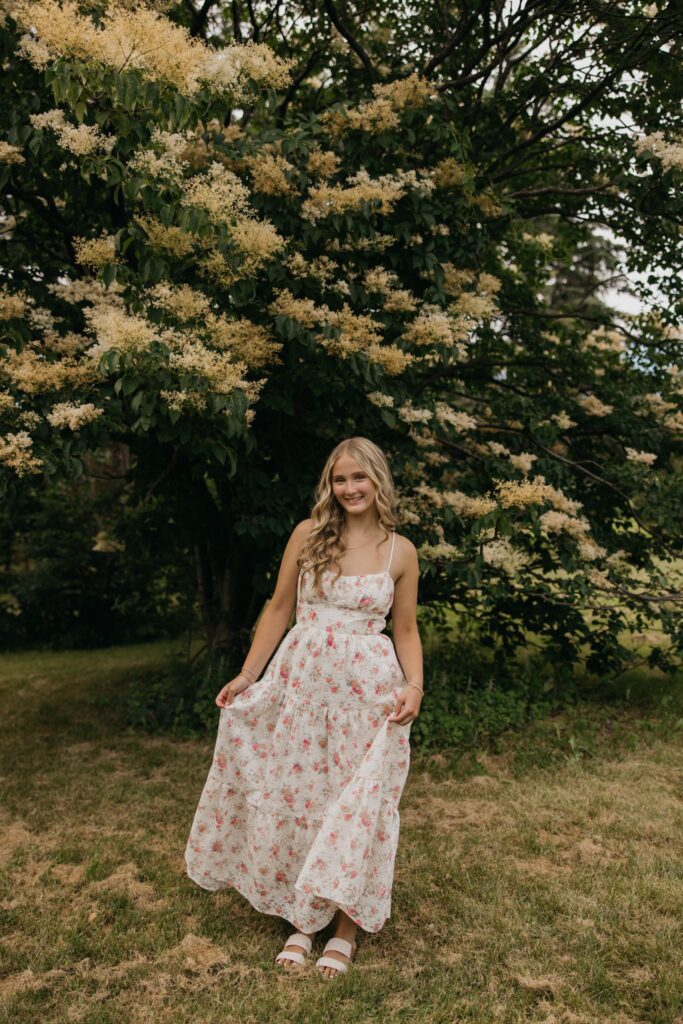 Senior girl in a floral maxi dress walking through grass beneath a blooming tree
