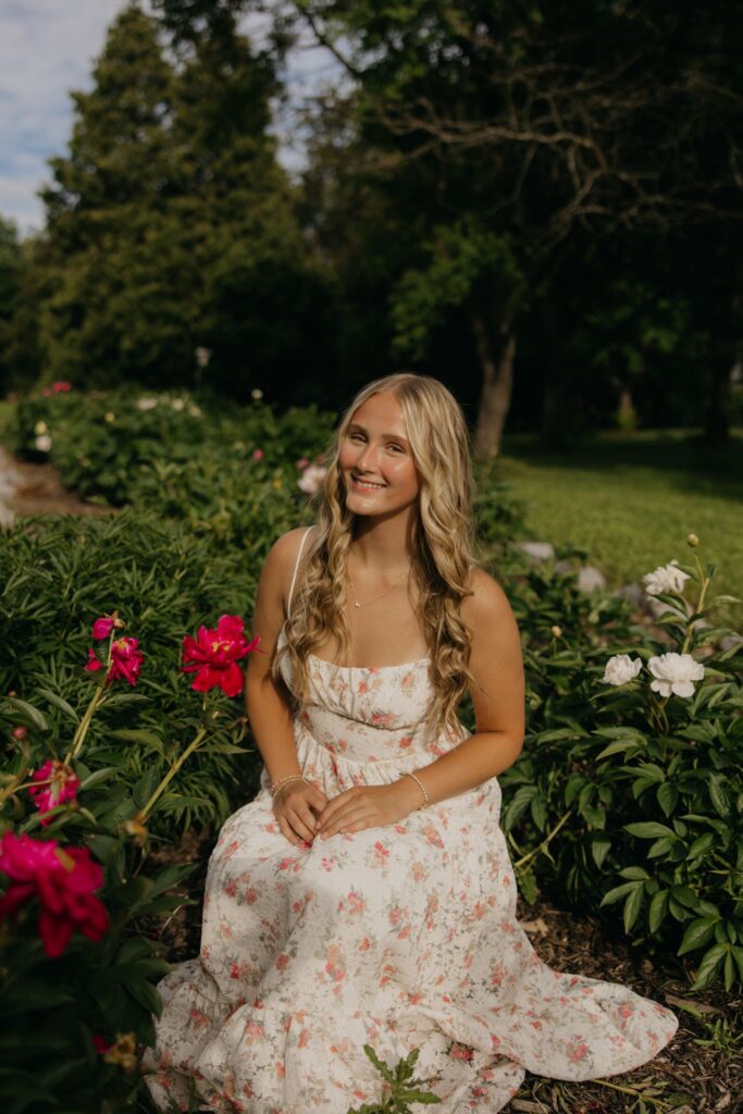 Senior girl in a floral maxi dress sitting among peonies and garden flowers