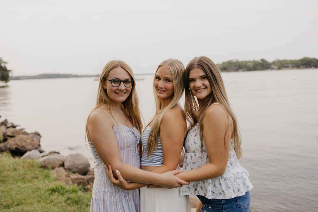 three friends smiling at the camera with arms linked by the water