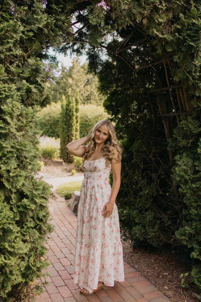 Senior girl in a floral maxi dress walking along a brick path under a garden archway