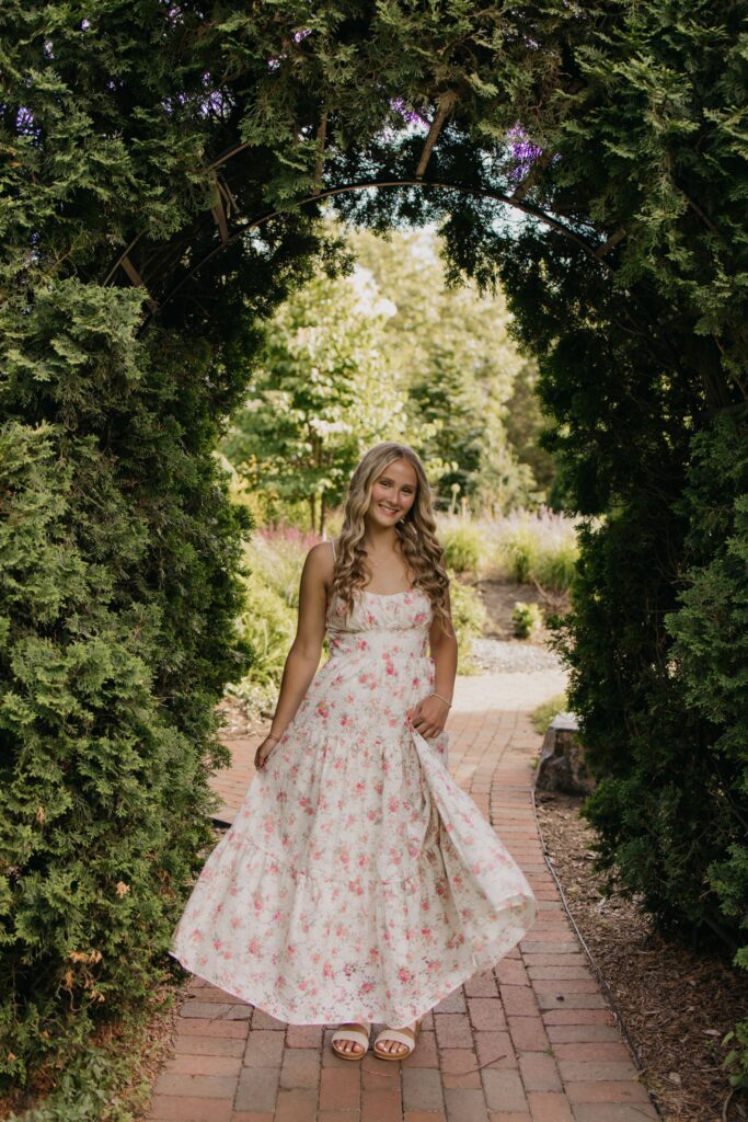 Senior girl in a floral maxi dress twirling under a garden arch on a brick pathway