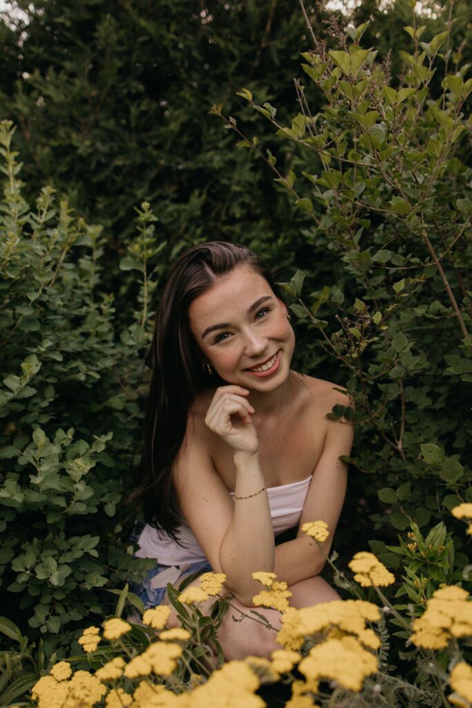 Senior girl sitting among yellow flowers resting her chin on her hand with a soft smile