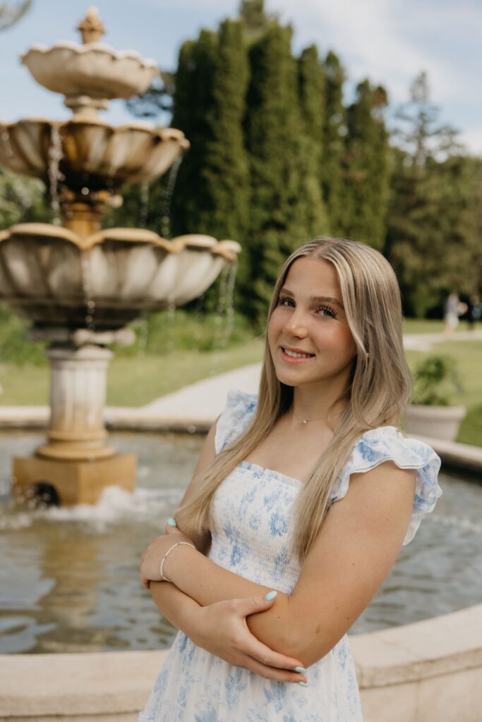 Senior girl in a blue floral dress standing beside a stone garden fountain