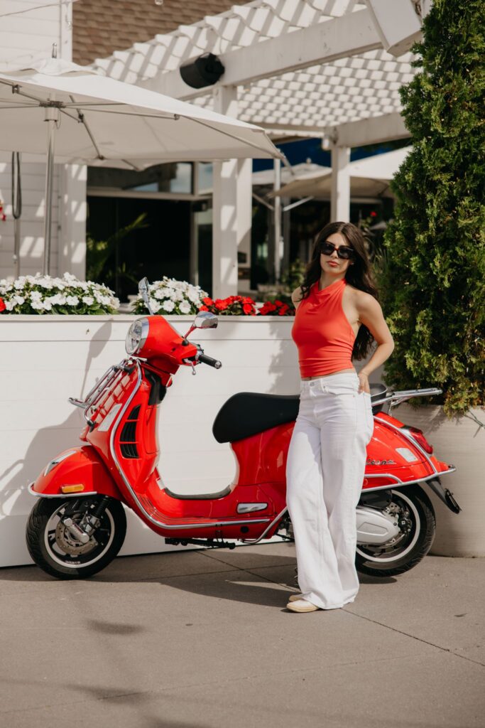 Senior girl in a red top and white pants standing beside a red Vespa scooter