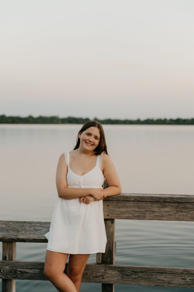 Senior girl in a white dress leaning on a wooden dock railing at sunset