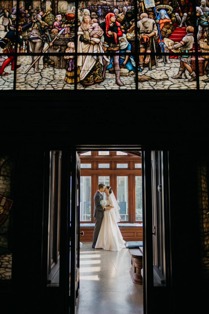 Stained glass window above a dim wood-paneled hallway with the bride and groom embracing in the distance at the American Swedish Institute