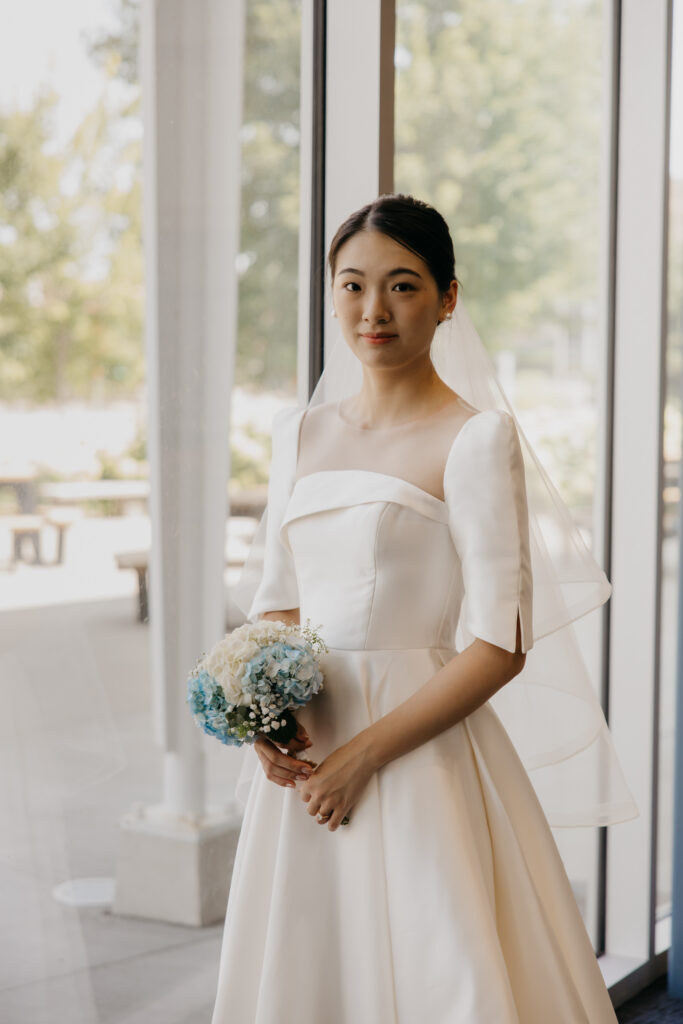 Bride poses alone holding her bouquet in front of floor-to-ceiling windows at the American Swedish Institute