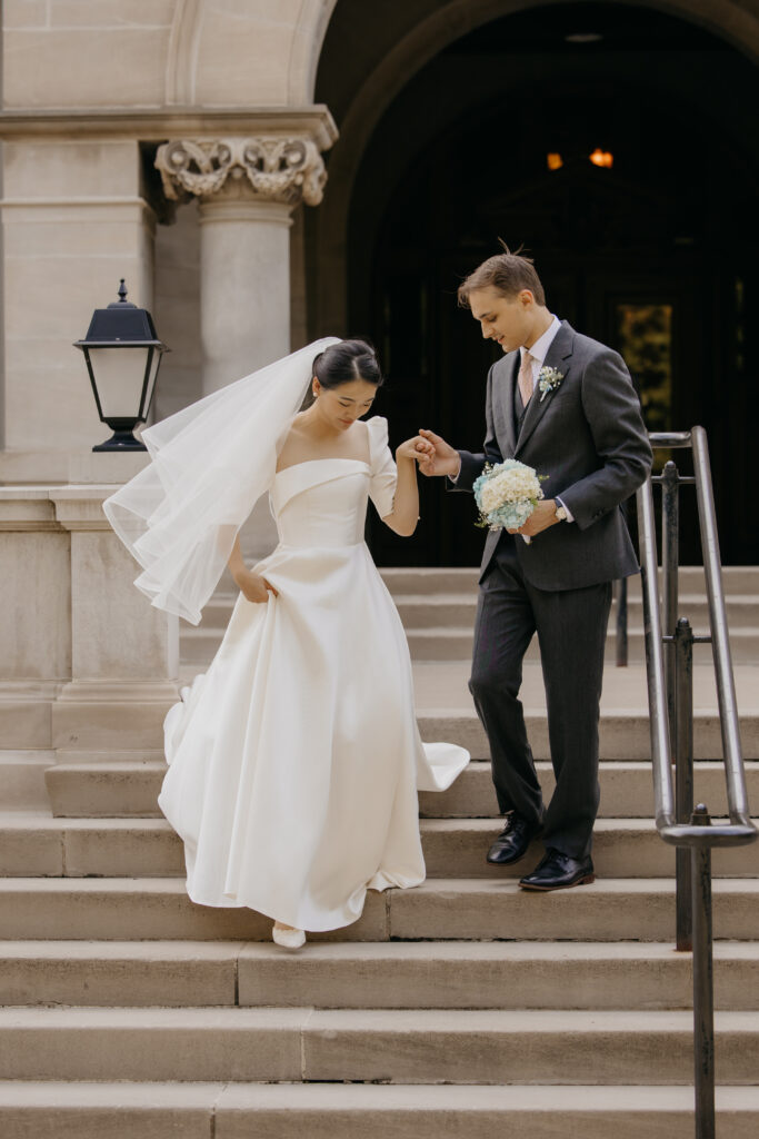 Bride lifts her gown while groom helps her down the stone steps outside Central Lutheran Church