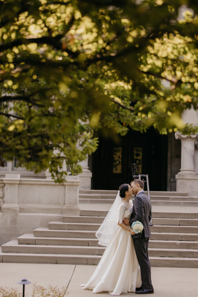 Bride and groom kiss on the church steps beneath a tree, framed by the stone columns of Central Lutheran Church