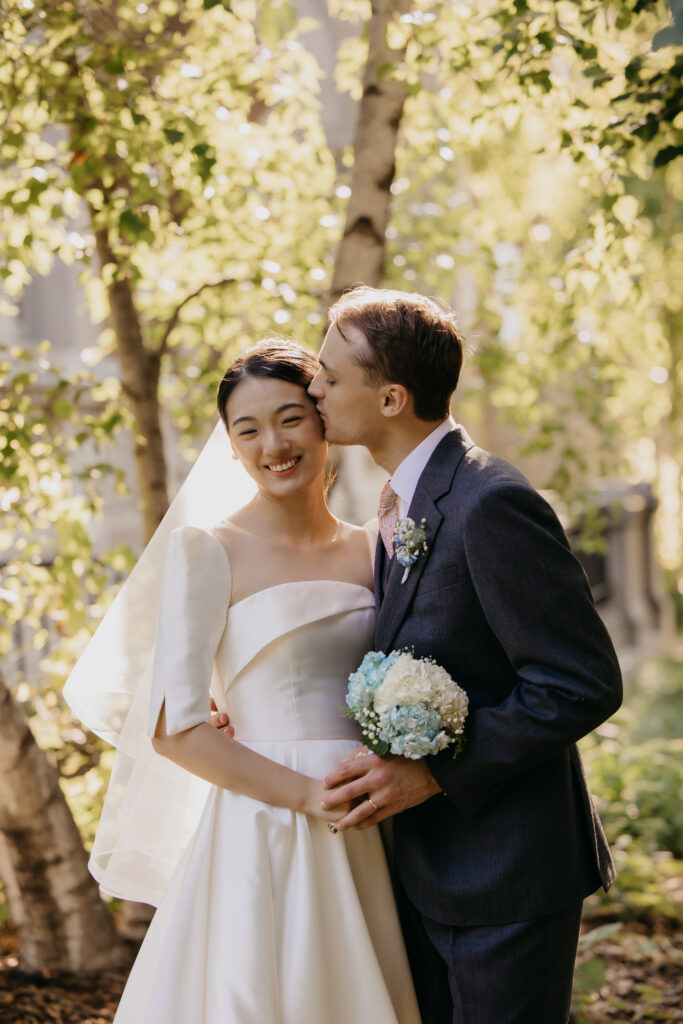 Groom kisses bride's cheek beneath dappled light in a birch tree grove at the American Swedish Institute gardens