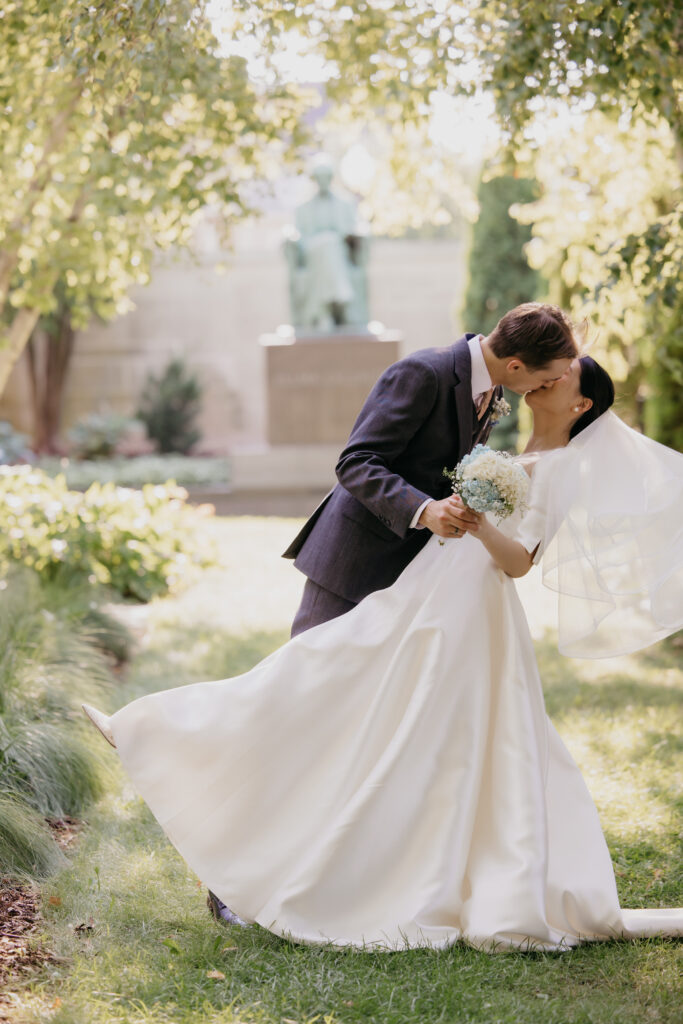 Groom dips bride for a kiss as her veil flows in the American Swedish Institute garden