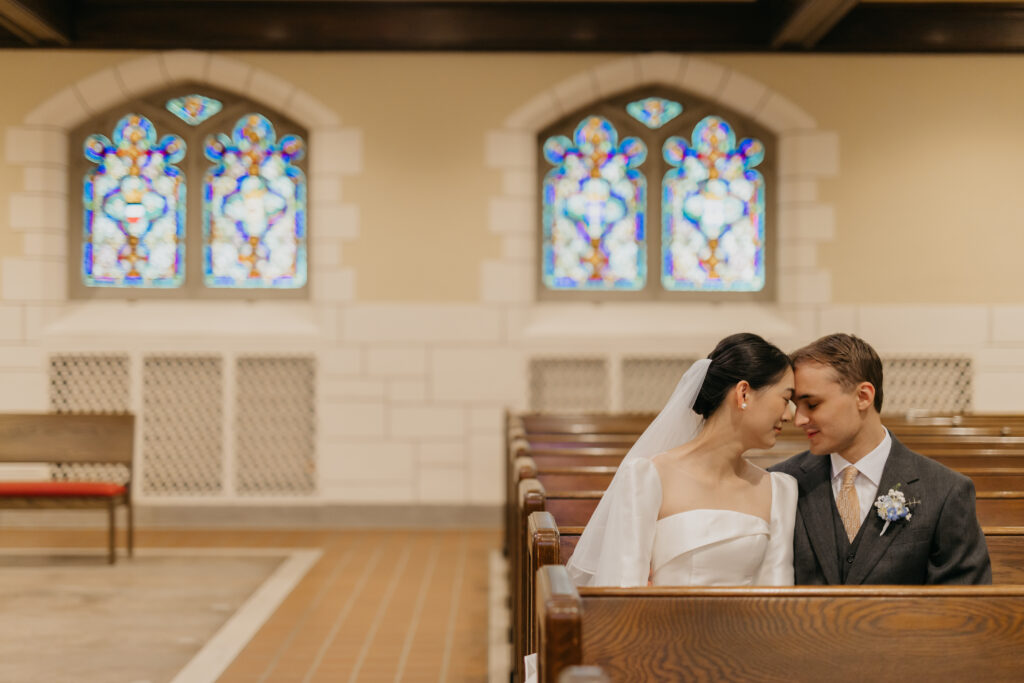 Bride and groom sit together in a wooden pew inside Central Lutheran Church near a stained glass window