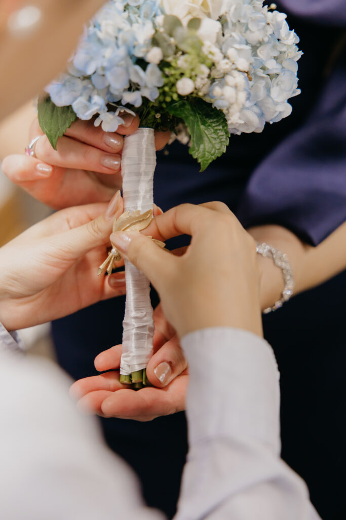 Close-up of hands wrapping ribbon around the blue and white hydrangea bouquet before the ceremony