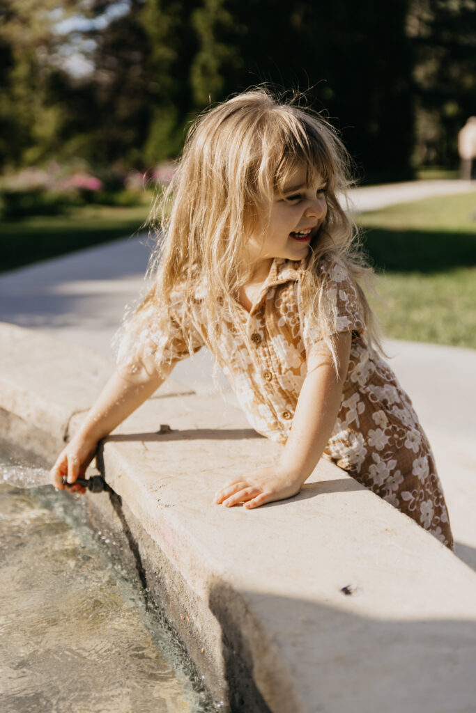 Young girl in a floral outfit leaning over a stone fountain ledge reaching toward the water