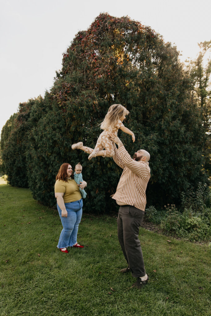Dad playfully tossing young daughter in the air on the lawn as mom holds baby and laughs