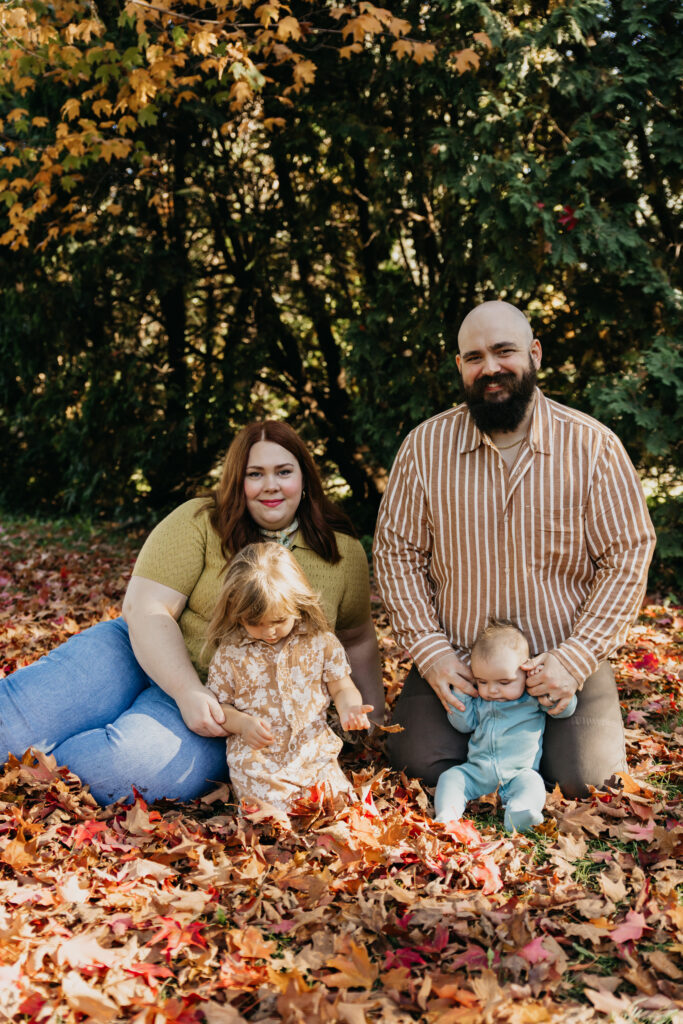 Family sitting together in a pile of fall leaves, dad holding baby and mom next to daughter in a floral outfit