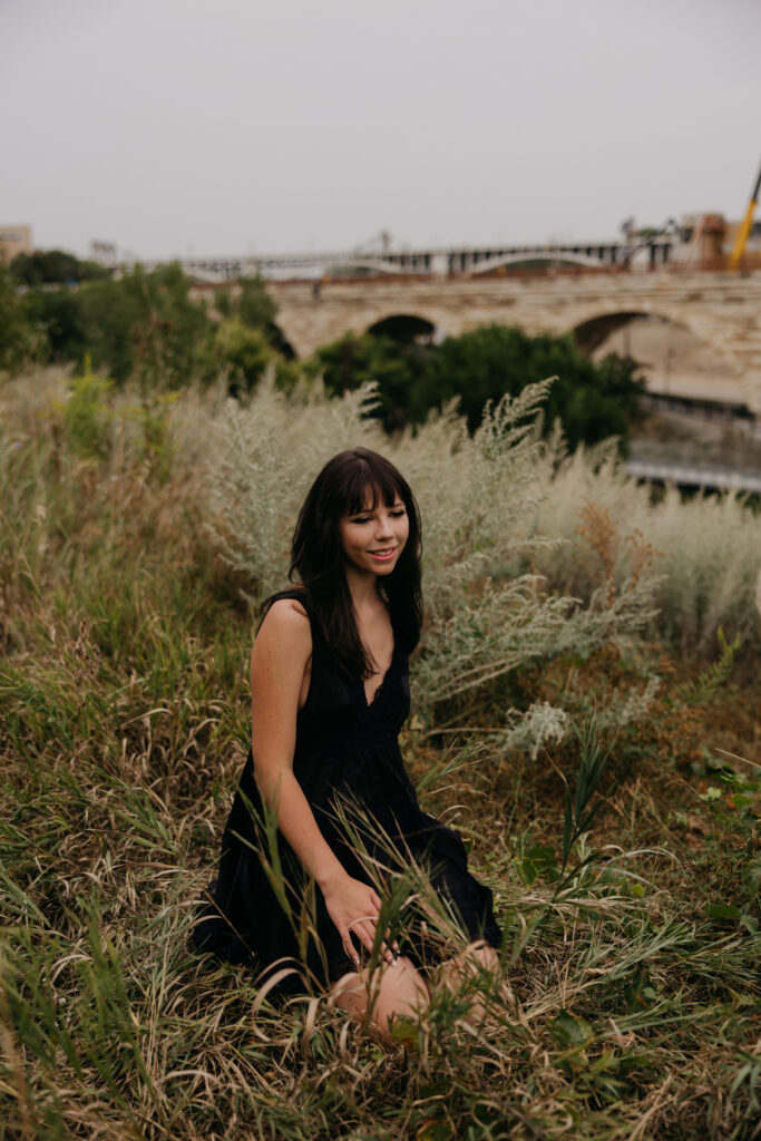 Senior girl kneeling in tall grass with the Stone Arch Bridge in the background