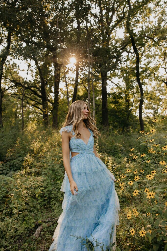Senior walking through backlit wildflowers in a blue ruffled dress at golden hour