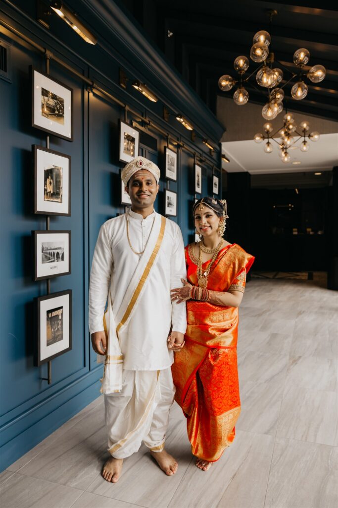 Groom in a white veshti and turban with his bride in an orange silk saree beside a navy gallery wall