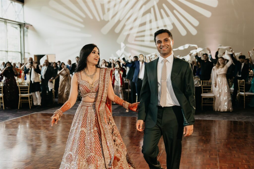 Bride in a gold lehenga and groom in a green suit making their reception entrance as guests wave napkins
