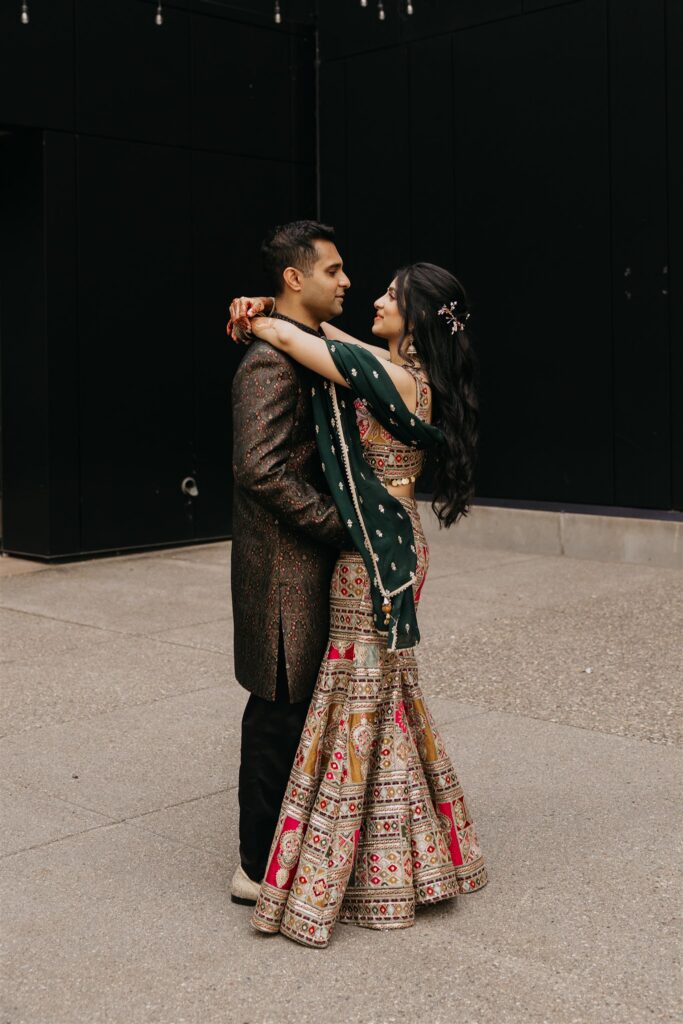 Bride in an embroidered lehenga and green dupatta embracing her groom against a dark wall