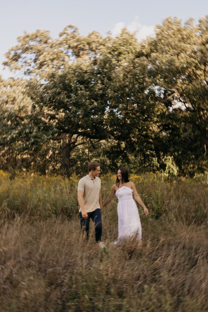 Wide view of a couple walking hand in hand through prairie grass and oak trees