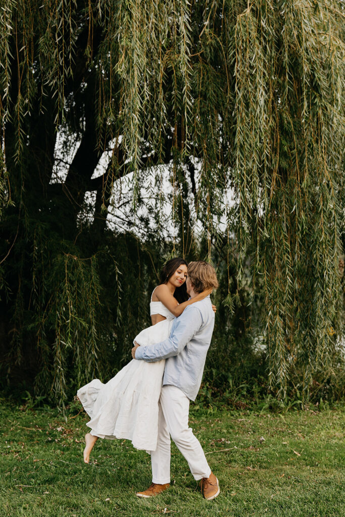Couple twirling on grass under a large weeping willow during their engagement session