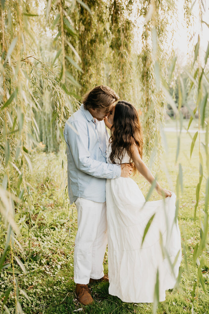 Couple kissing among willow branches, she in a white maxi skirt and he in light blue