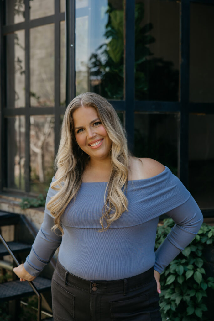 Caitlin Weseman smiling outdoors in an off-the-shoulder blue top in front of dark steel windows