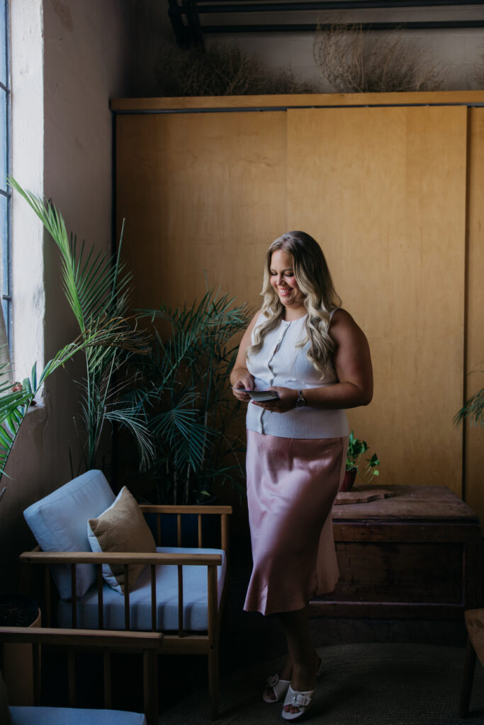 Caitlin Weseman standing by a sunny window with plants in a white top and pink skirt