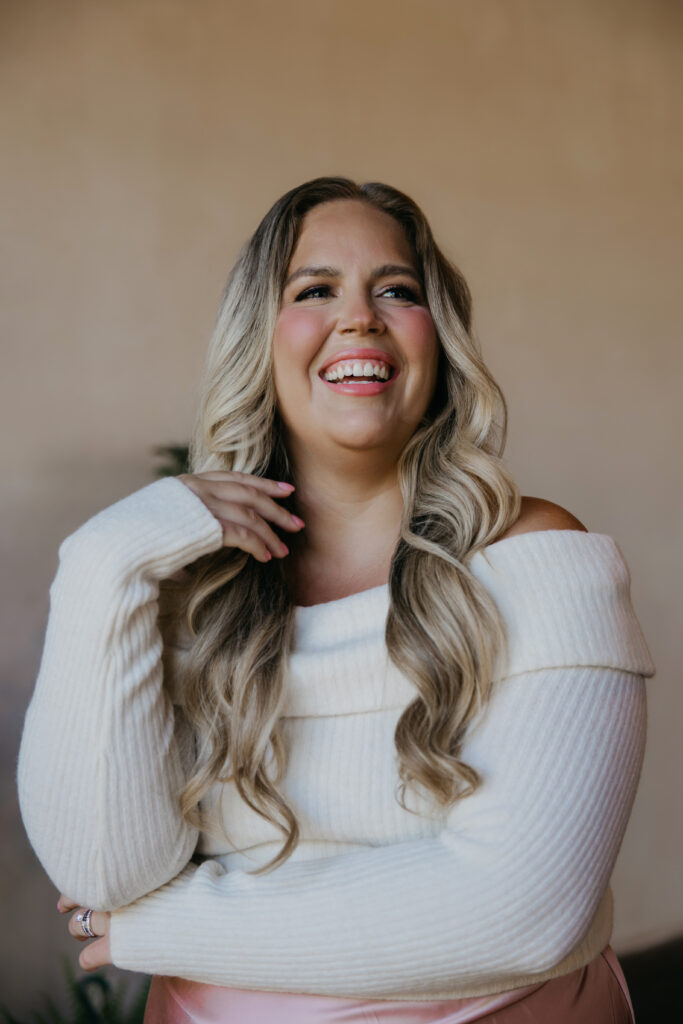 Caitlin Weseman laughing in a cream off-the-shoulder sweater, a warm astrologer headshot