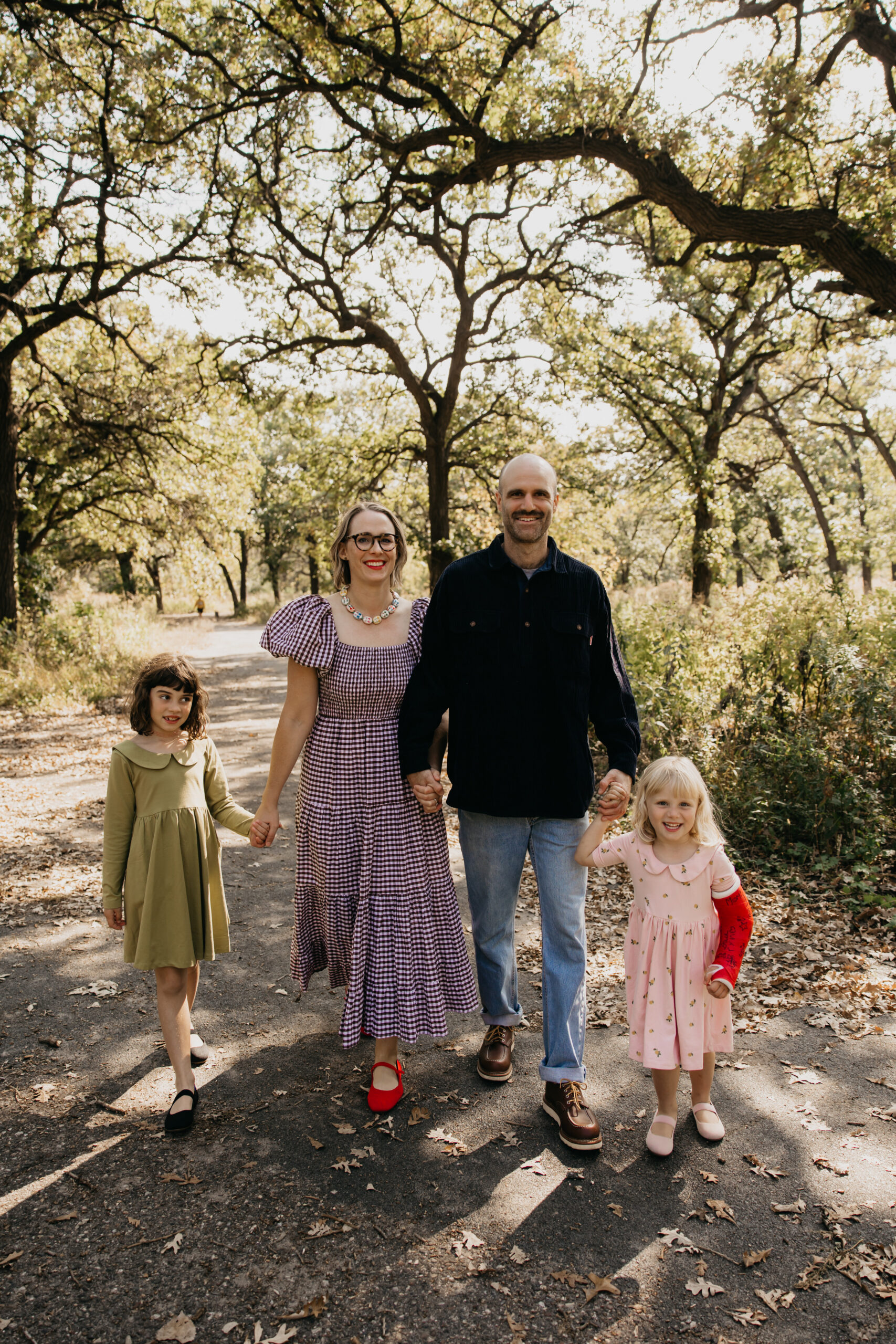 Family of four holding hands while walking down a sunlit wooded path during a fall family photo session