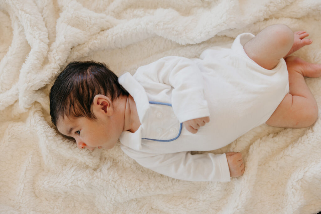 Newborn in a white collared romper with blue trim lies on a cream sherpa blanket, eyes open.