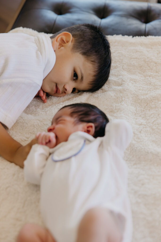 Big brother lies face to face with his sleeping newborn sibling on a cream blanket, a simple newborn and sibling pose.