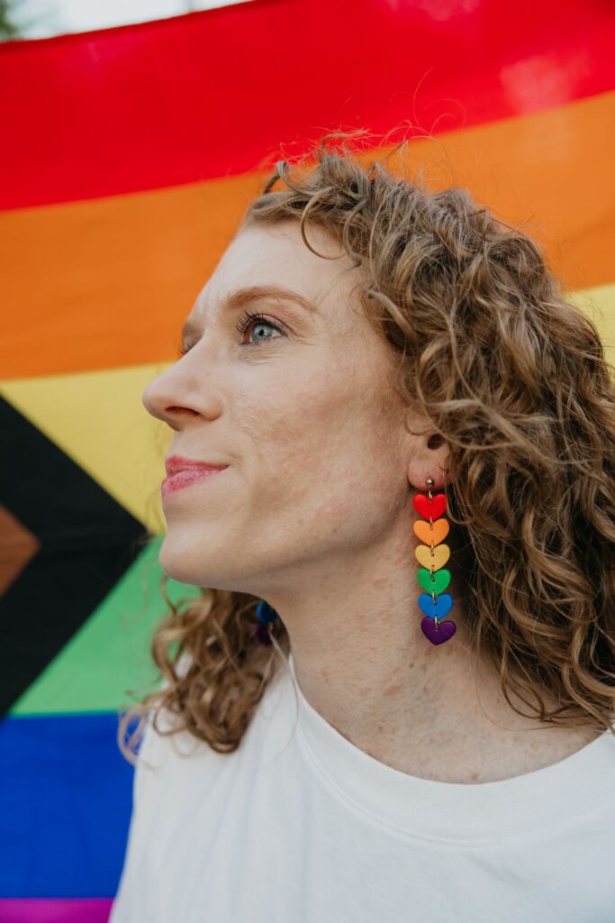 Woman with curly hair and rainbow heart earrings posed in front of a Progress Pride flag