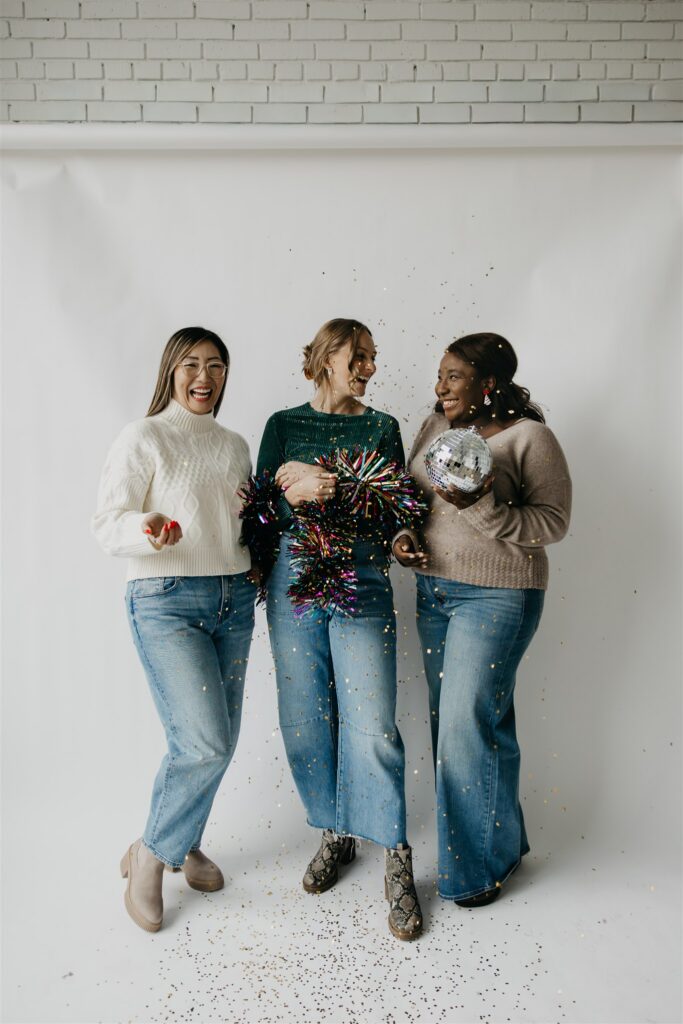 Three women in sweaters and jeans laughing together with tinsel and a mirrored disco ball