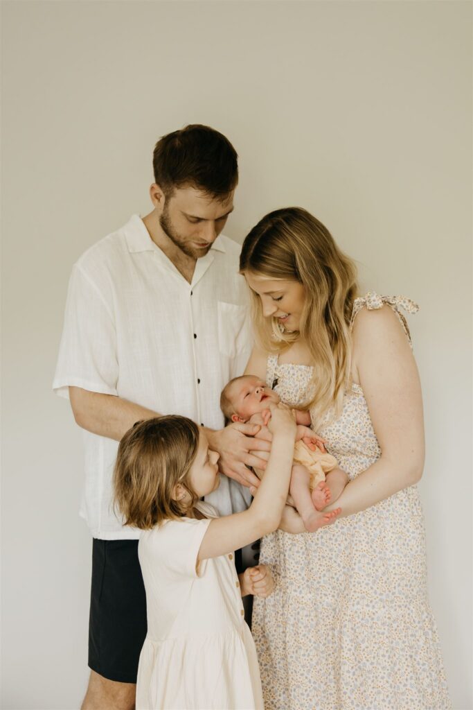 Parents stand against a white wall holding their newborn while their young daughter reaches up to touch the baby.