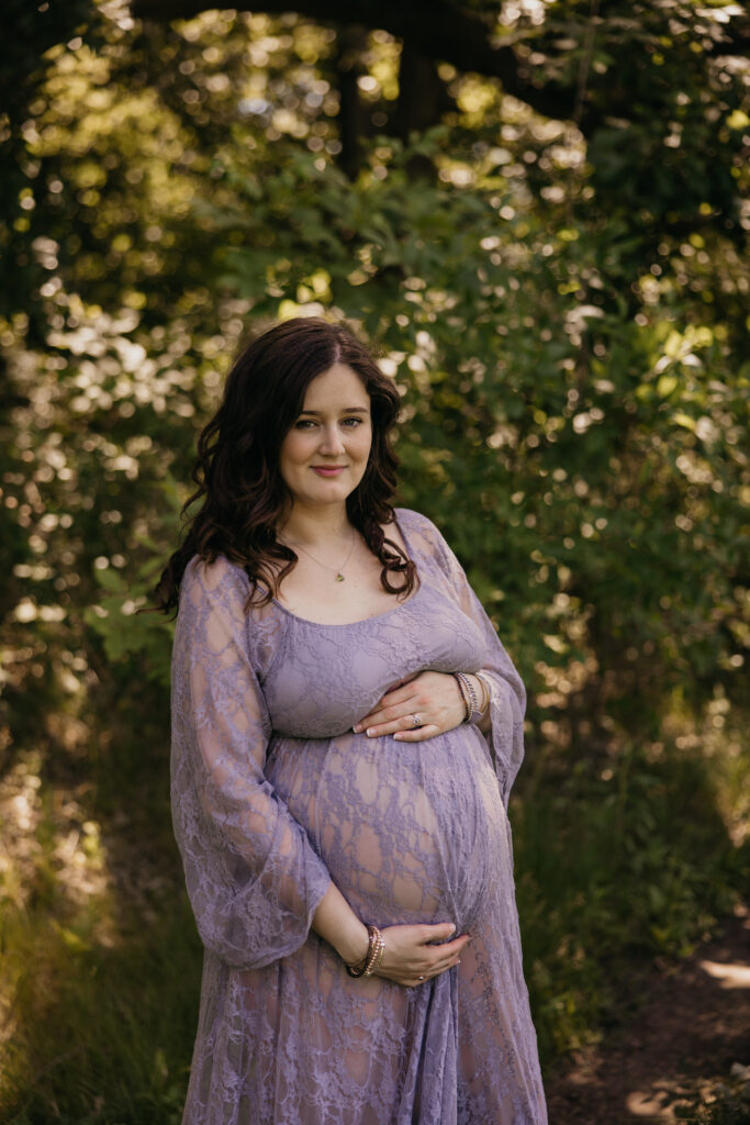 Pregnant woman in flowing lace maternity dress standing in dappled forest light
