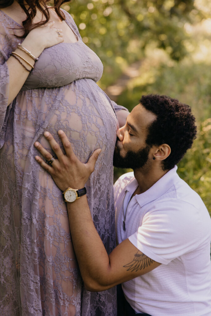 Close-up of a man kissing his partner's bare belly beneath a sheer lace maternity dress